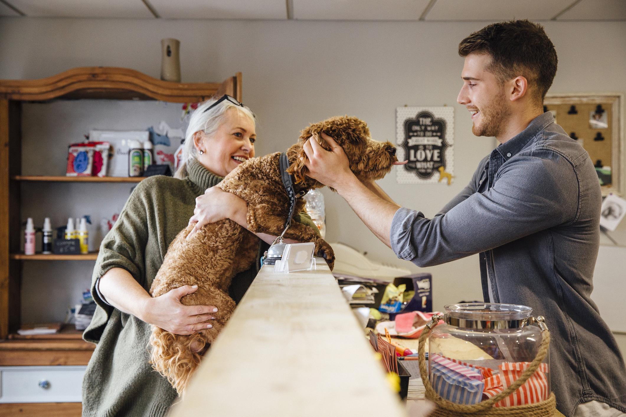 A woman holds up her dog to a man behind the counter, the man is petting the dog on their ears