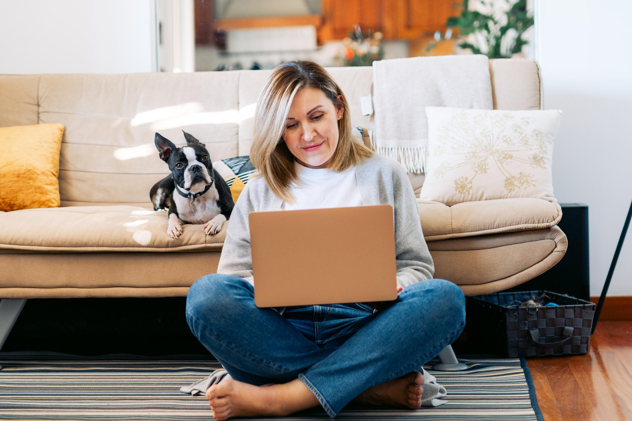 A woman sits with a laptop in her lap cross-legged against her sofa, her Boston Terrier lies next to her on the sofa