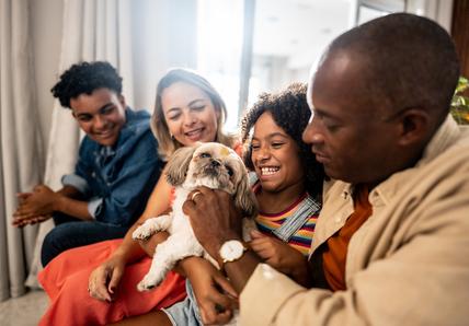 A family of four sits on a couch, smiling and playing with a small dog.