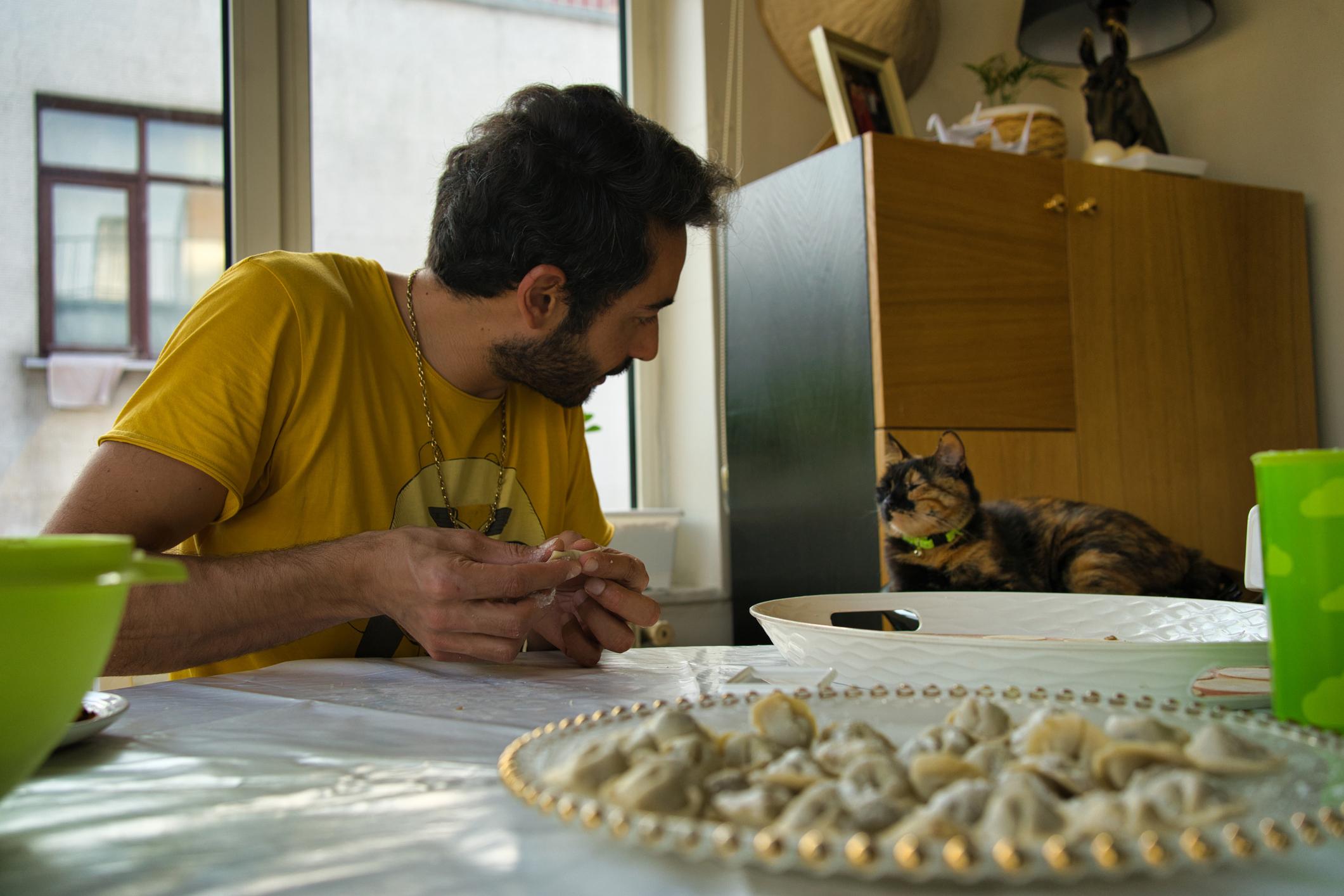 A man in a yellow shirt makes pasta at a table while looking at a calico cat lying near a cabinet.