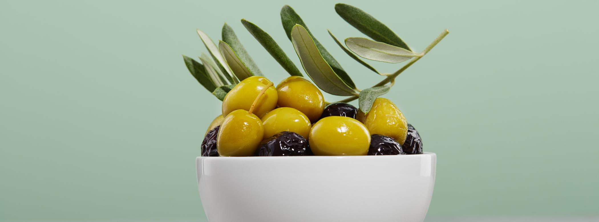 A white bowl filled with green and black olives and olive branches, set against a light green background.