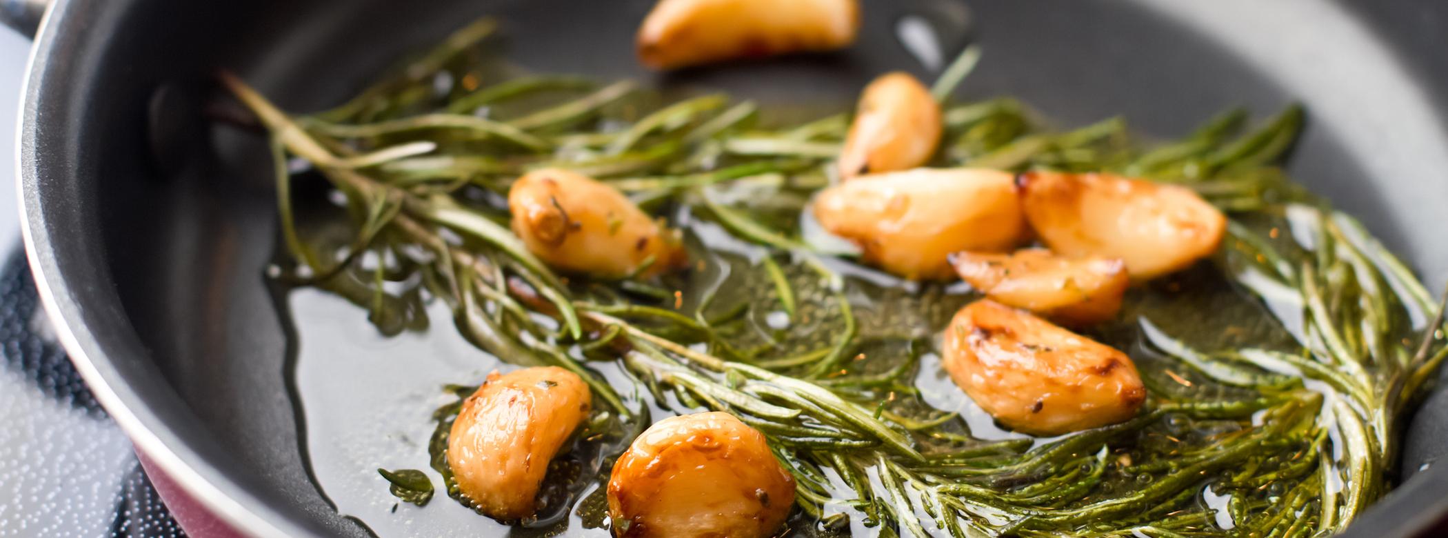 Garlic cloves and rosemary sprigs sizzling in oil in a frying pan.