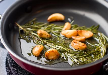 Garlic cloves and rosemary sprigs sizzling in oil in a frying pan.