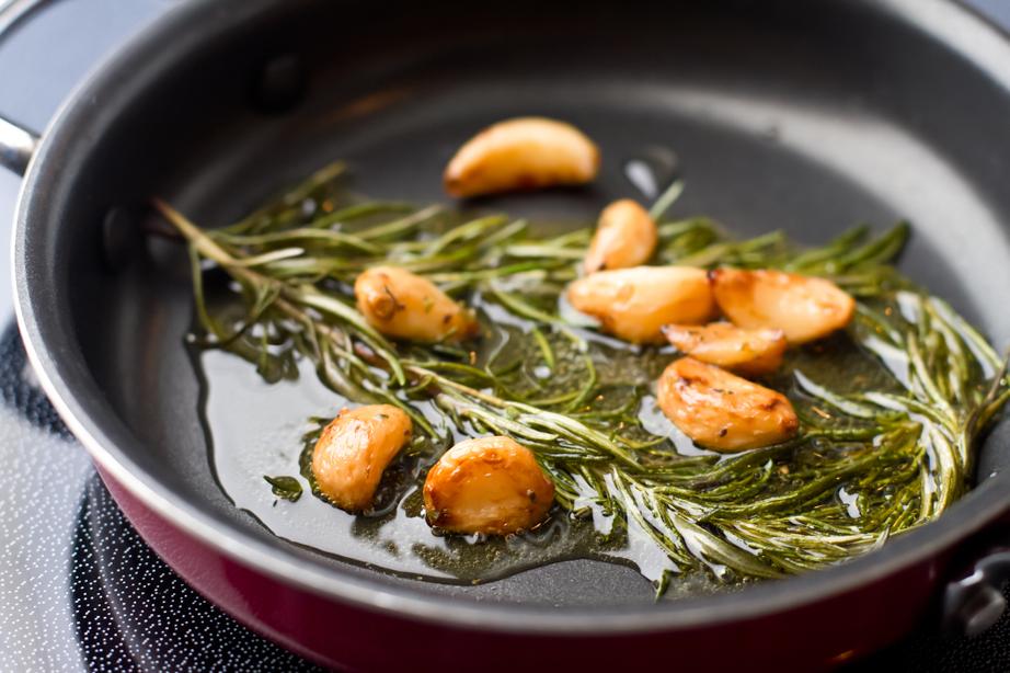 Garlic cloves and rosemary sprigs sizzling in oil in a frying pan.