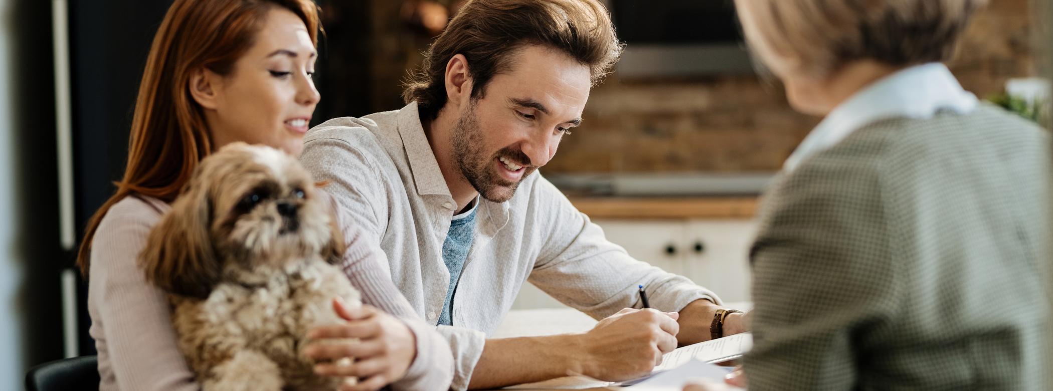 A couple sits at a table with a fluffy dog, while a professional woman, seen from the back, talks to them. The man is signing papers.