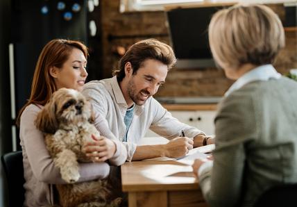 A couple sits at a table with a fluffy dog, while a professional woman, seen from the back, talks to them. The man is signing papers.
