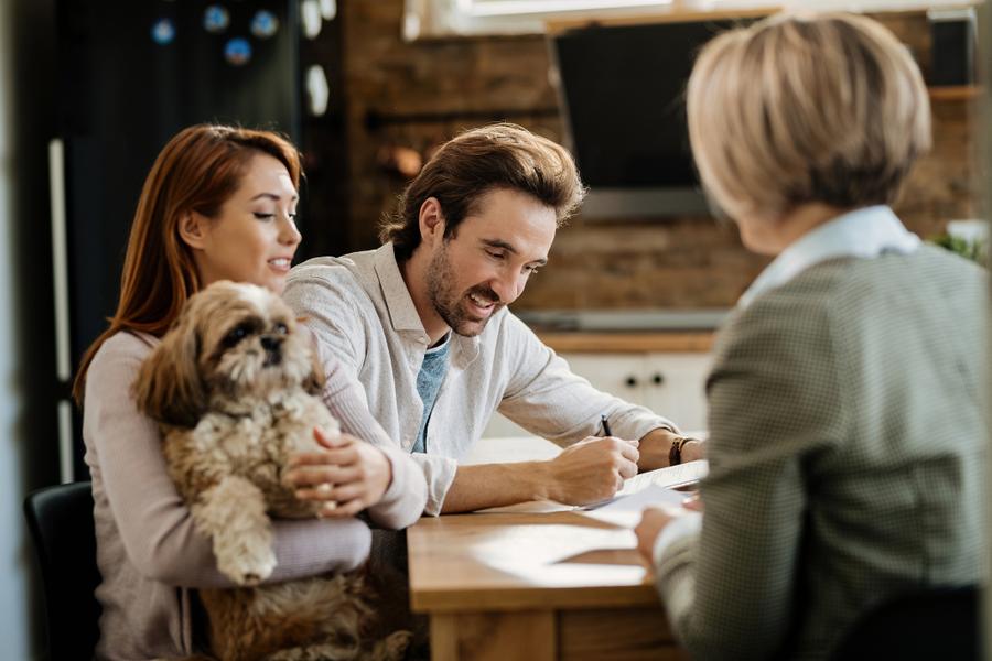 A couple sits at a table with a fluffy dog, while a professional woman, seen from the back, talks to them. The man is signing papers.