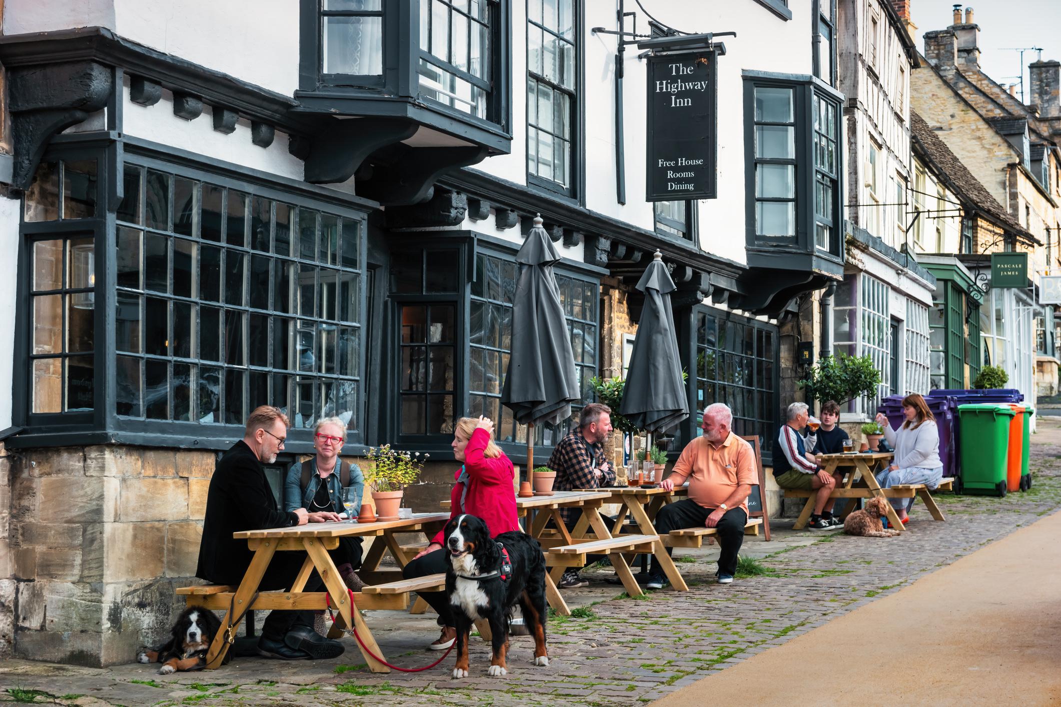 People and dogs sitting outside a historic inn with large windows, enjoying drinks at wooden tables on a cobblestone street.