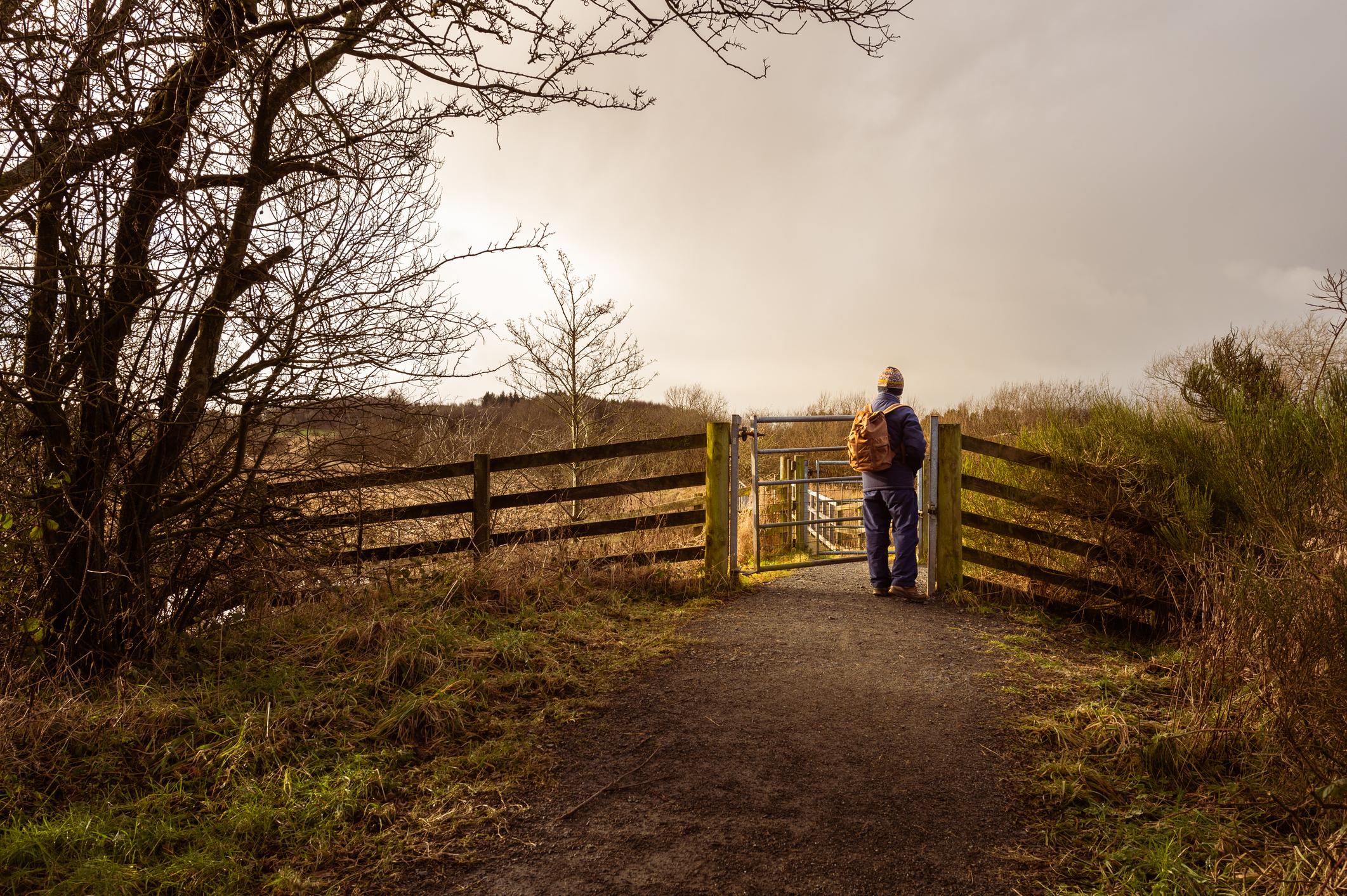 A man stands near a gate on a pathway in the Dumfries, Scotland - the day is overcast with the sun struggling to get through the clouds