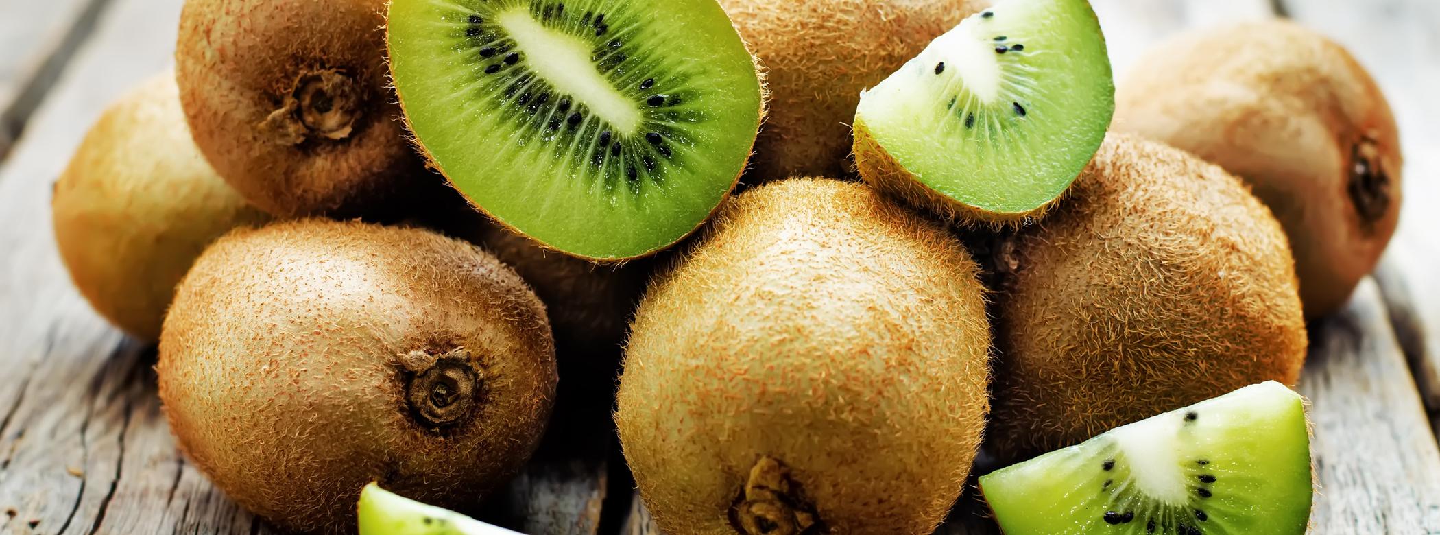 A pile of whole kiwis with a few slices showing vibrant green flesh and black seeds, set on a rustic wooden surface.