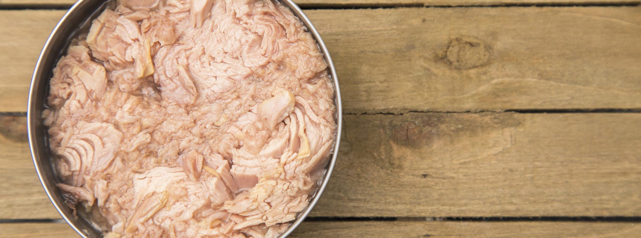 Open can of tuna on a wooden table, showing pink flaked fish inside.