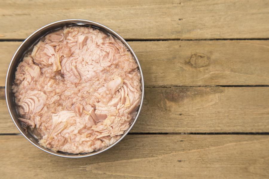 Open can of tuna on a wooden table, showing pink flaked fish inside.