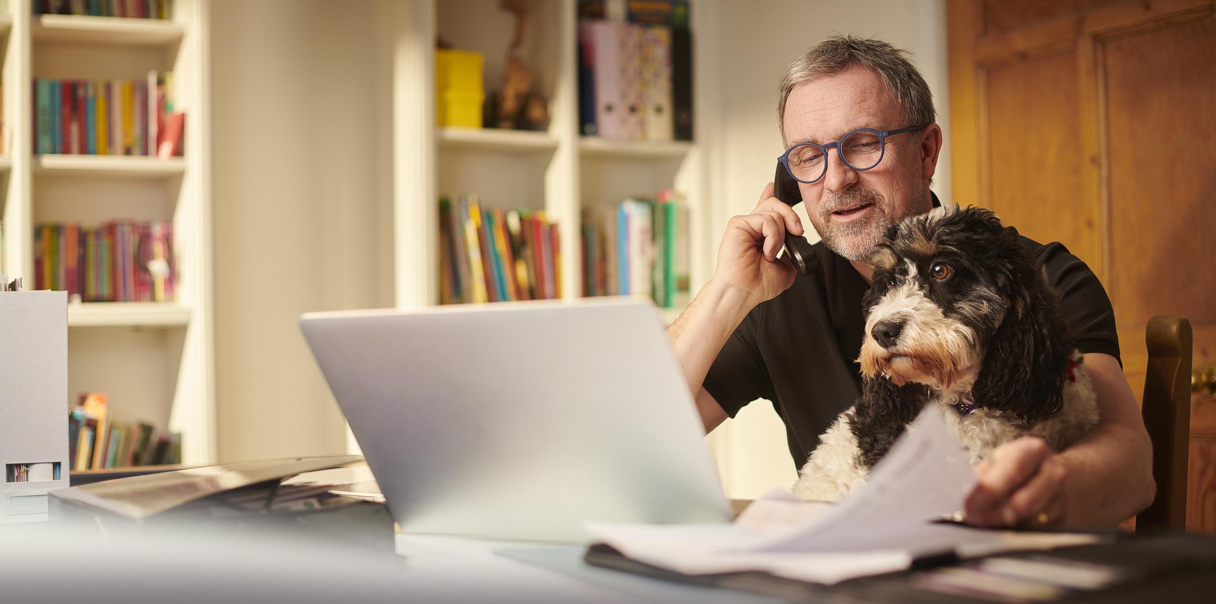 A man talks on the phone while using his laptop, his black-and-white dog sits on his lap
