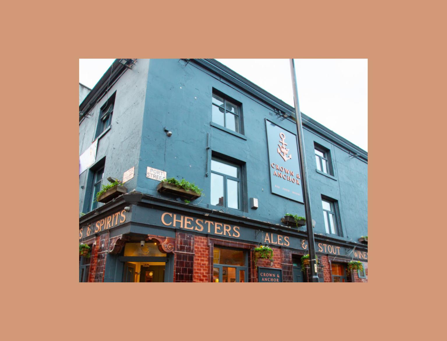 A corner pub painted blue - with a sign saying "Crown &amp; Anchor" on the facing side. It has a banner wrapping around it, listing Ales, Stout, Wines &amp; Spirits.