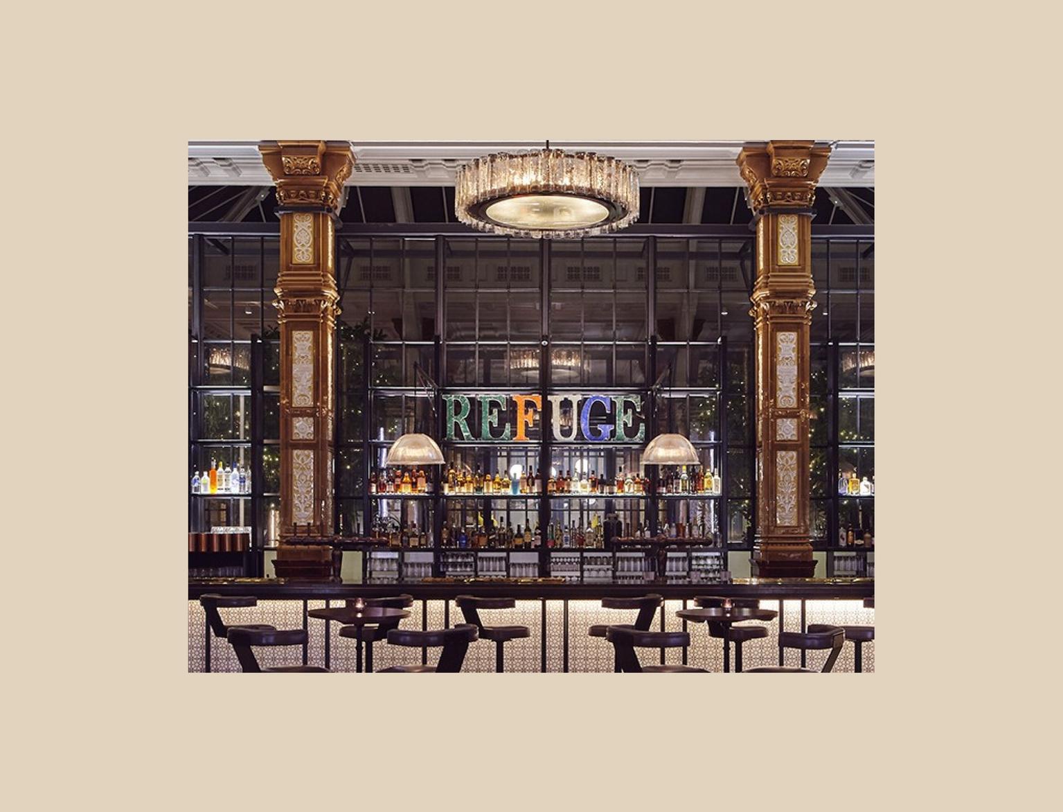 Opulent bar interior with ornate golden columns and intricate tiled pillars, featuring a well-lit bar counter, plush seating, and a 'REFUGE' neon sign in the background.