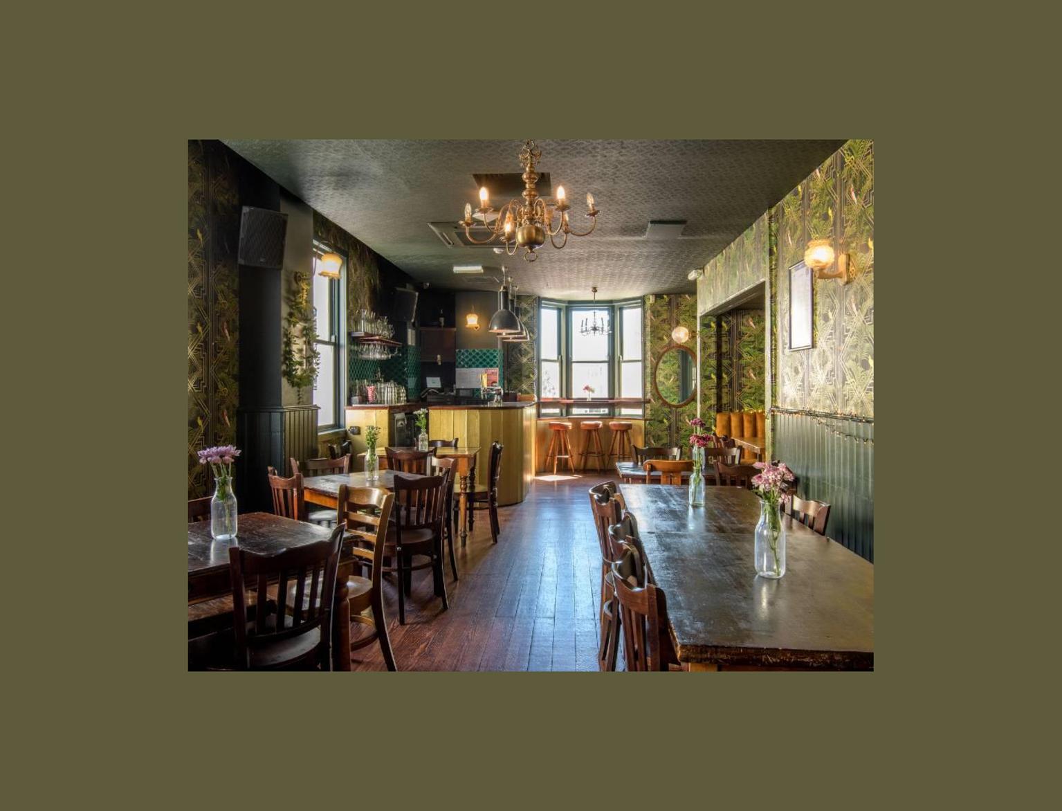 Inside of a bar with green patterned wallpaper, wooden tables and chairs.