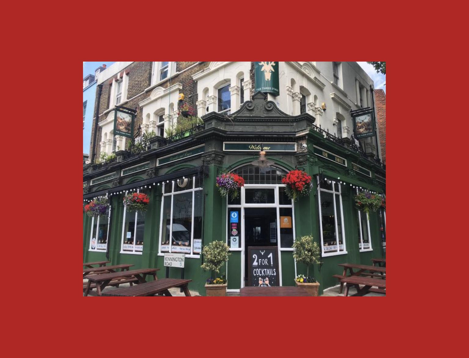 Traditional British pub on a corner, with vibrant flower baskets and '2 for 1 Cocktails' offer. The green facade stands out under a clear sky, inviting guests to its outdoor seating area.