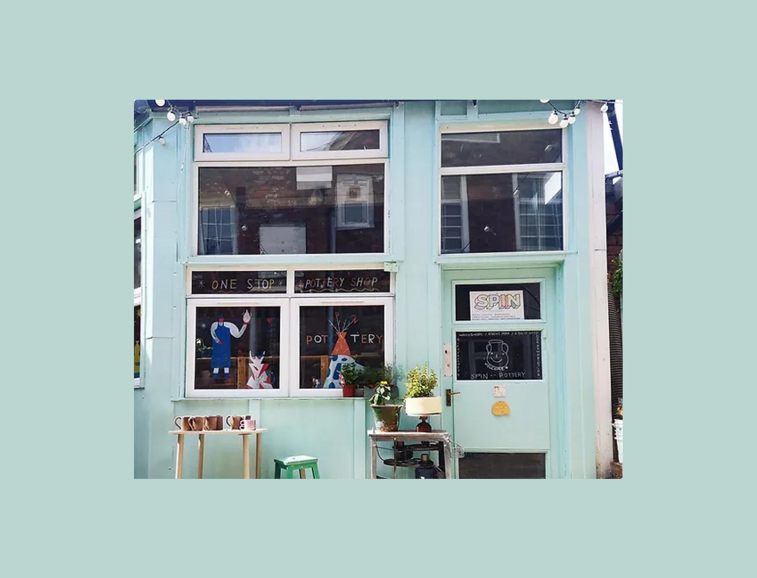 Spin Pottery Shop's pale blue facade features large windows displaying pottery and creative signs, inviting a warm, artistic vibe.