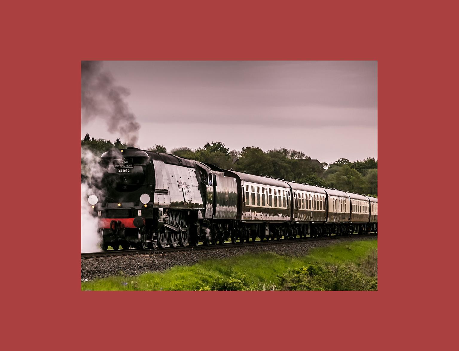 A vintage steam locomotive moves along the tracks, emitting a cloud of steam against a lush, tree-lined background.