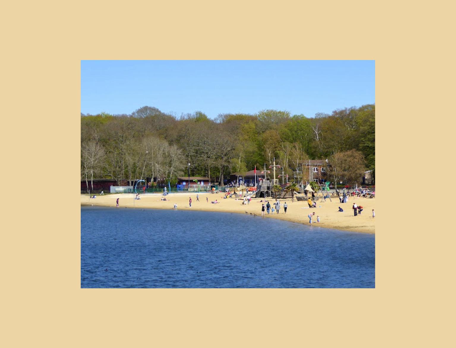 Scenic view of Ruislip Lido's sandy beach and playground bustling with families enjoying a sunny day by the water, surrounded by a leafy forest