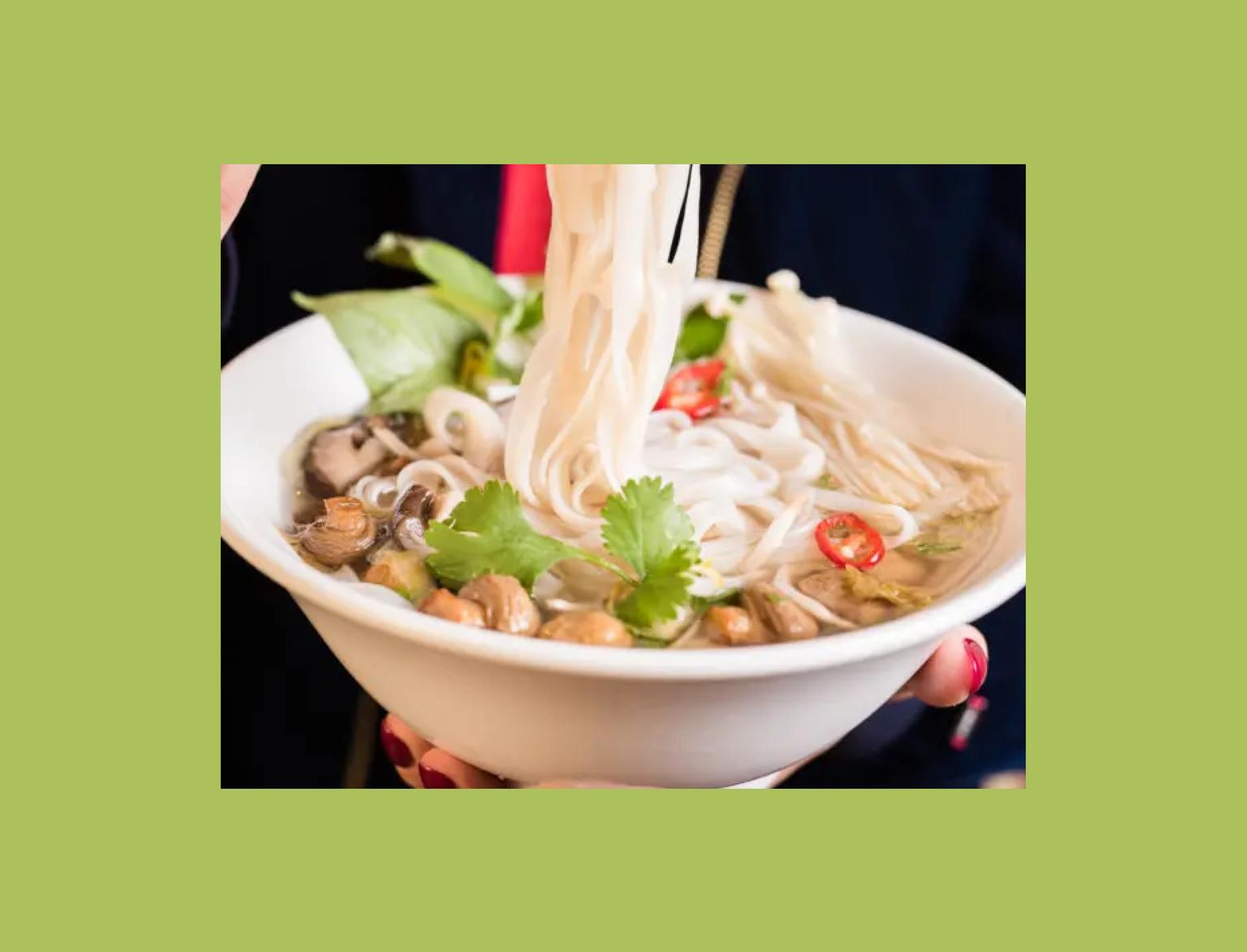 A person holds a bowl of pho, using chopsticks to lift noodles. Fresh coriander, mushrooms, and sliced chilies top this vibrant and steamy soup.