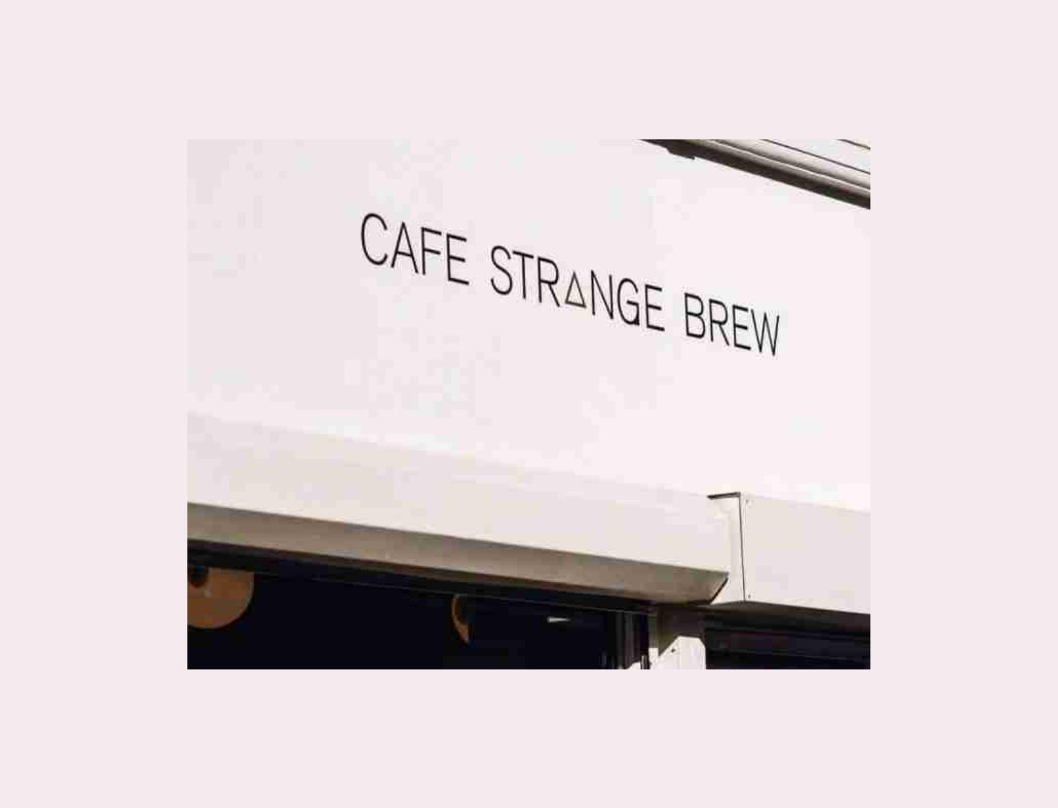 Outside Cafe Strange Brew, people wait in line under a sign. Sunlit, casual scene with plants at the cafe entrance.