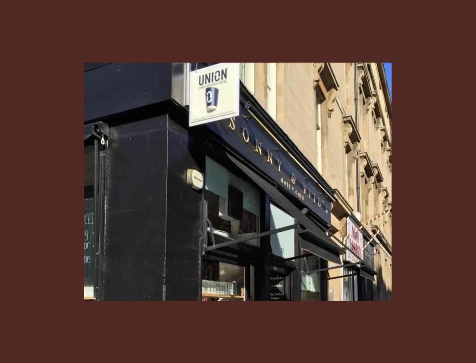 Exterior of a cafe on a corner, featuring outdoor seating with round tables, brown chairs, and a black facade under a sunny sky.