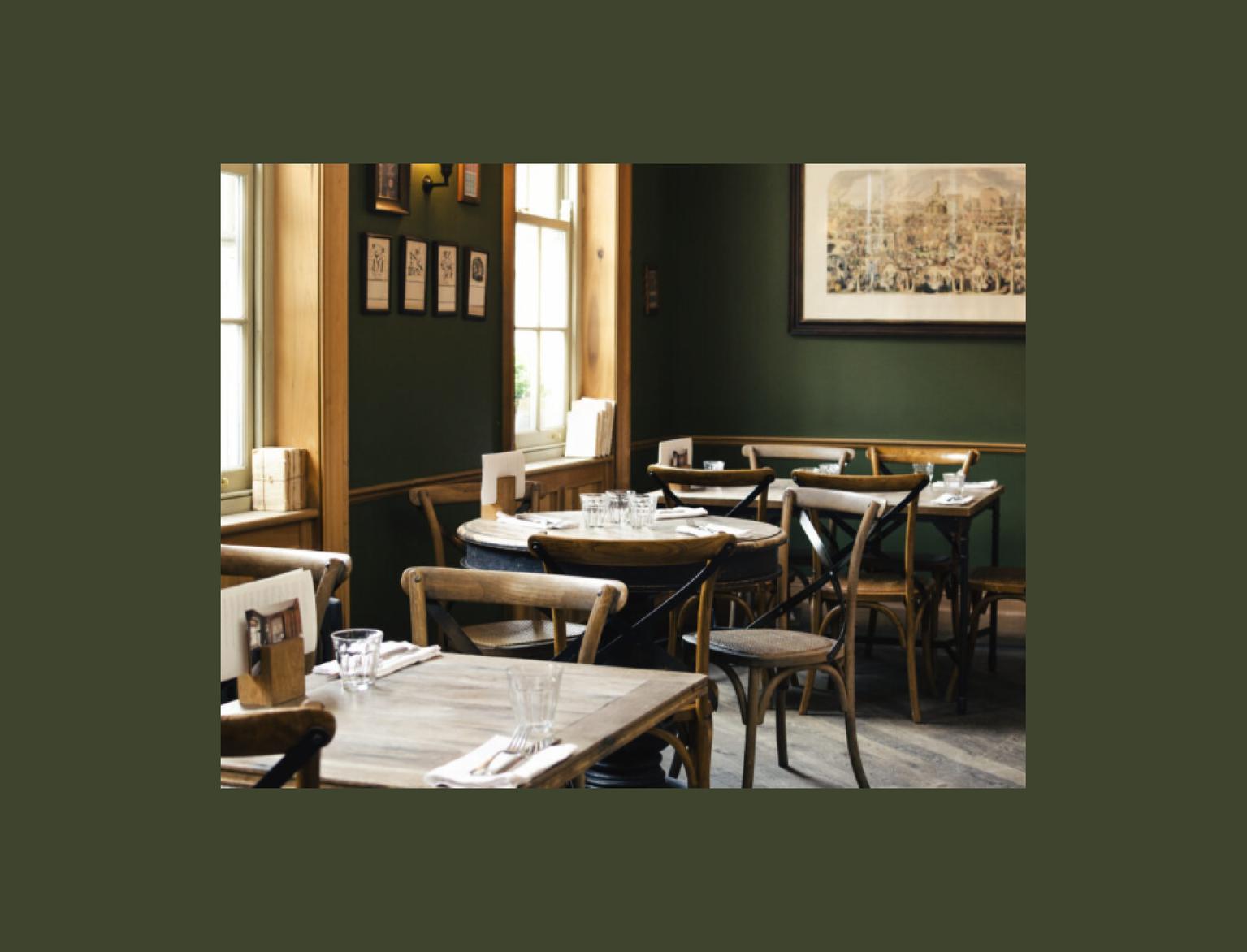 Intimate pub setting with dark green walls and wooden furniture. Natural light highlights the classic bentwood chairs and rustic tables set for dining. The walls are adorned with framed pictures and a large vintage-style painting.