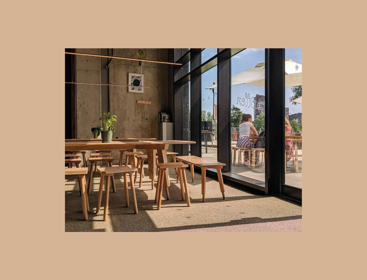 Inside a café with concrete walls, large glass windows, wooden tables, and stools; patrons sit near the entrance, enjoying the sunny view outside.