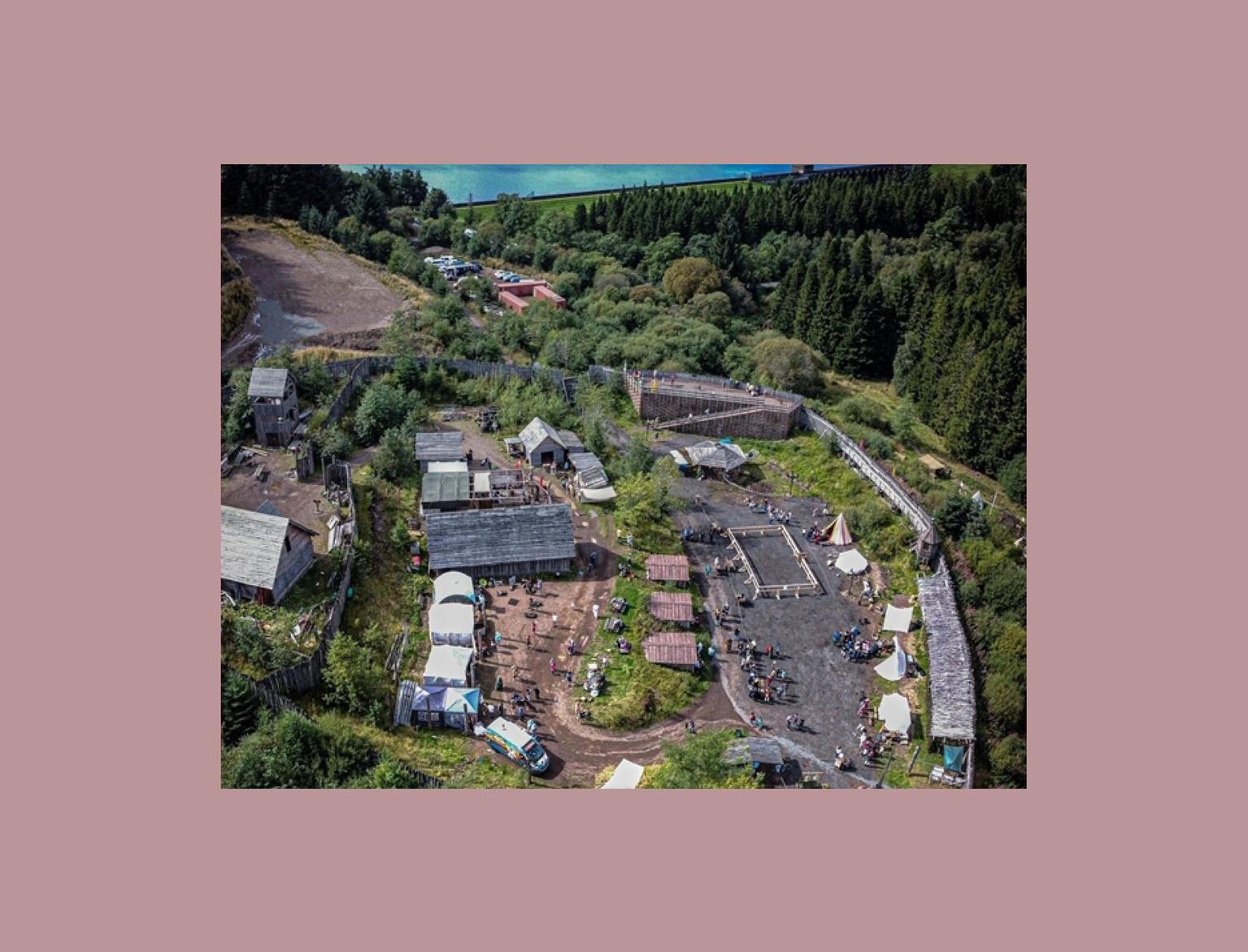 An aerial view of Duncarron Medieval Village with clustered buildings and tents, set against a backdrop of lush forests and a serene lake.