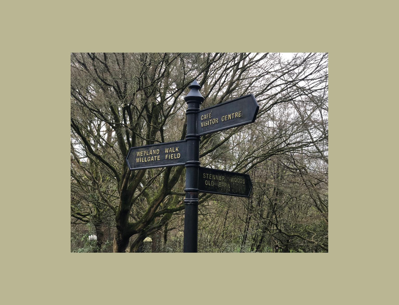 A directional signpost with arrows pointing to various locations, including "Wetland Walk", "Millgate Field","Cafe", "Visitor Centre", "Stenner Woods", and "Old Barn", set against a backdrop of leafless trees.