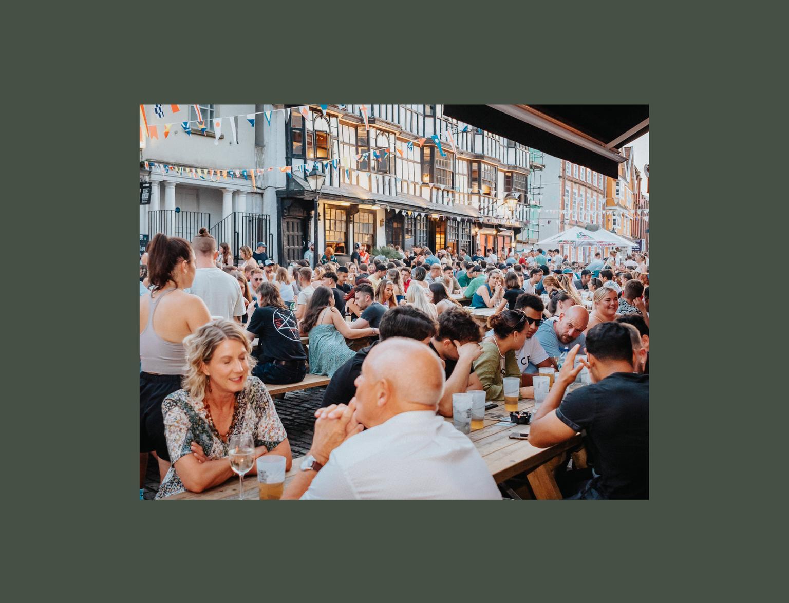 Lots of people sat drinking on pub benches in busy street