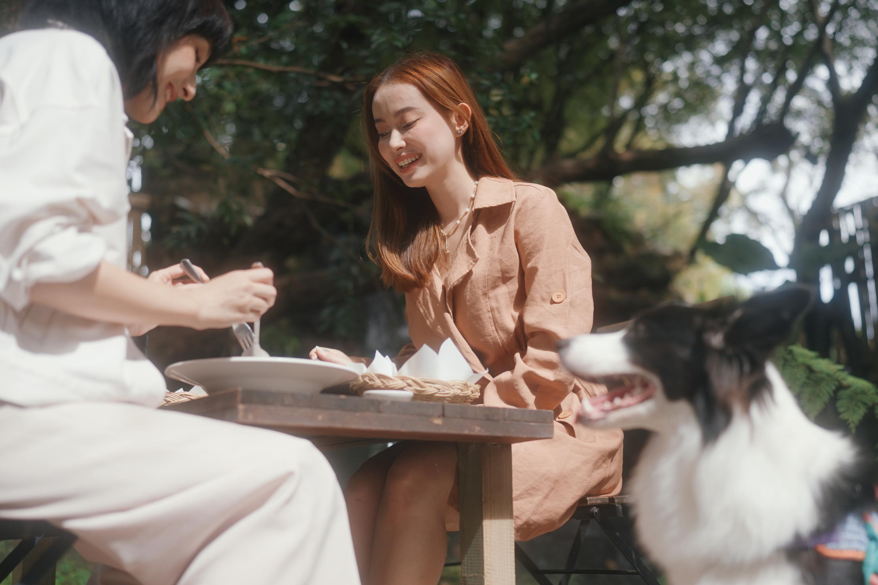 Two women sitting at an outdoor table enjoying a meal, with a dog sitting nearby in a lush, green setting.