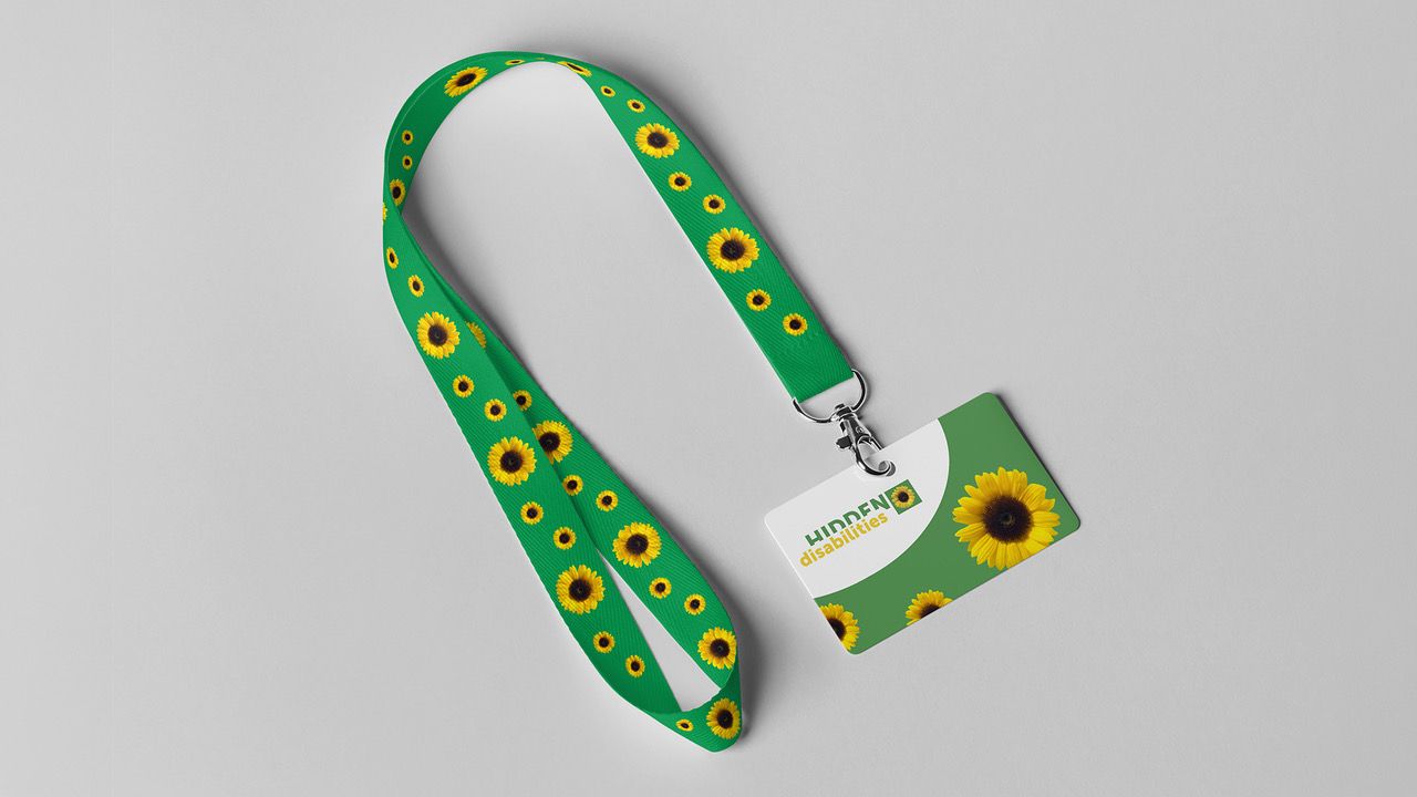 Hilft im ÖV die unsichtbare Behinderung sichtbar zu machen: Der Sunflower Lanyard. Bild: Silvia Meierhofer Grüner Bändel mit Sonnenblumen bestickt. Eine weiss grüne Karte mit Sonnenblumen und dem Schriftzug "Hidden disabilities" ist an den Bändel gehängt.