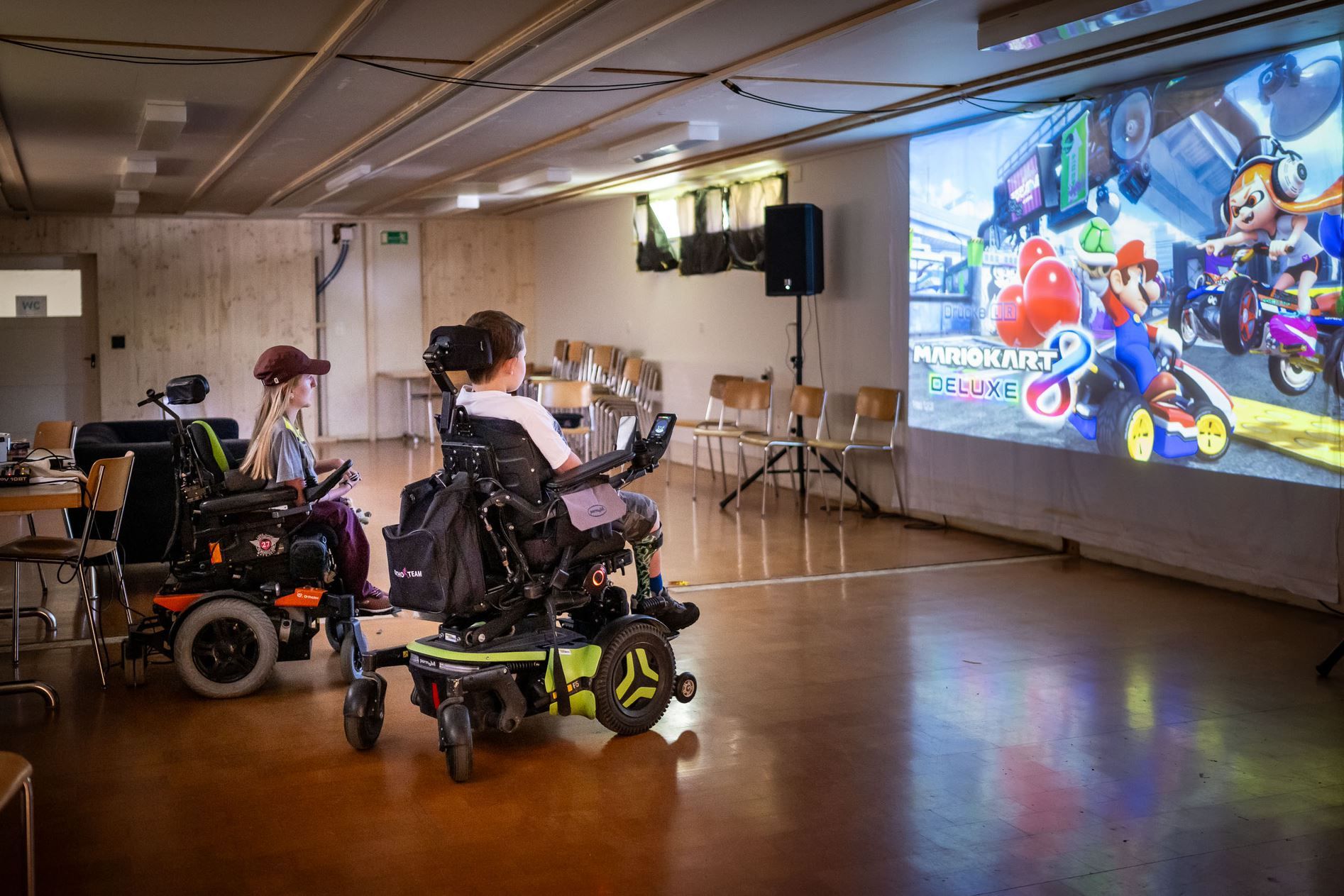 Zwei Rollstuhlfahrer am Gamen. An der Wand hängt eine grosse Leinwand.