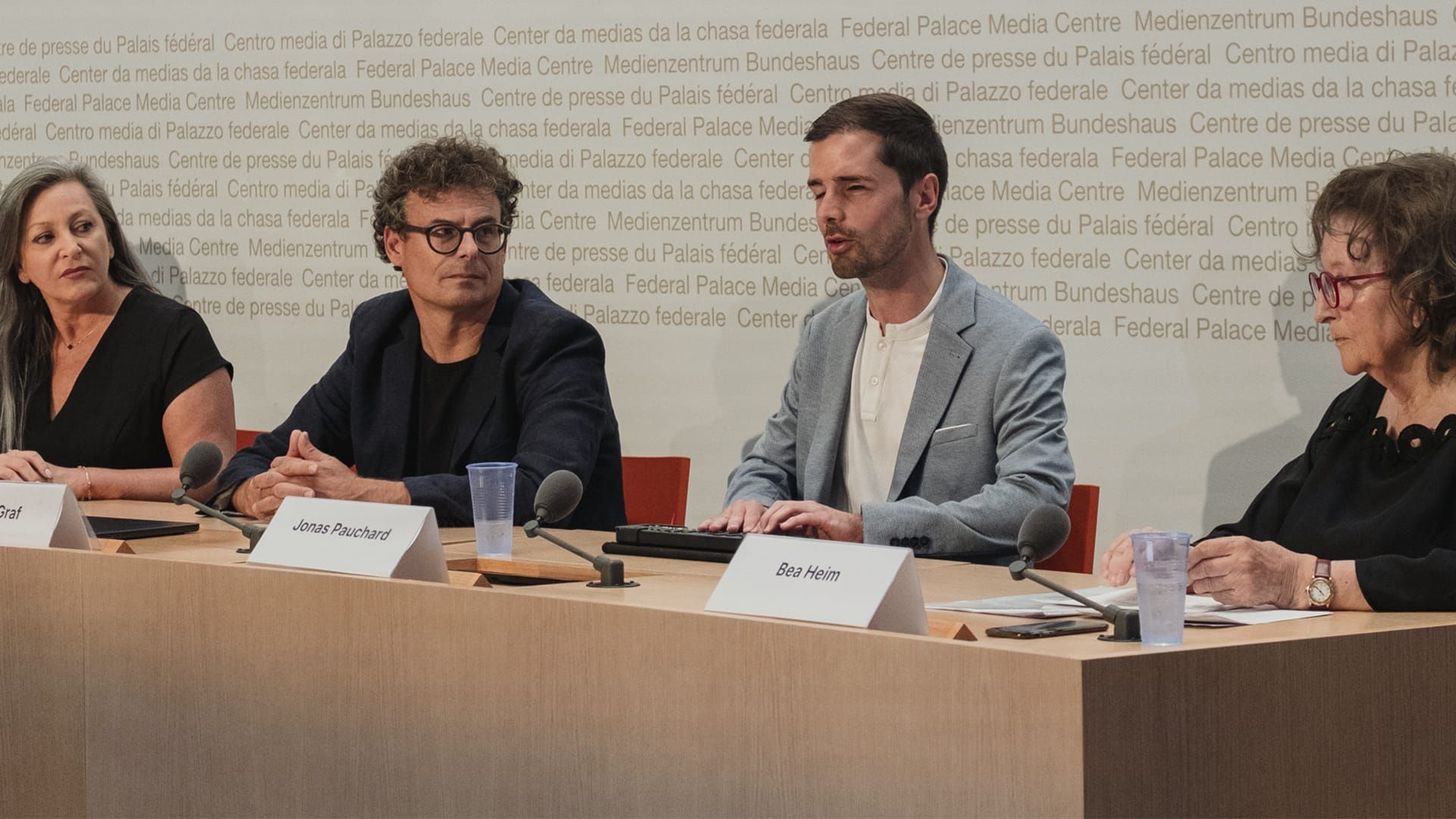 Jonas Pauchard (2 v.r.) spricht gemeinsam mit Daniel Graf, Bea Heim, Nuria Gorrite und weiteren Personen an der Medienkonferenz der Allianz pro E-ID vom 19. August 2025 im Medienzentrum des Bundeshauses Jonas Pauchard sitzt gemeinsam mit drei anderen Sprecher:innen an einem nur teilweise sichtbaren Pult. Er trägt kurz geschnittene braune Haare und ein graues Jackett über einem weissen Hemd. Auf dem Pult steht ein Mikrofon, im Hintergrund ist die Schrift des Medienzentrums des Bundeshauses sichtbar. Pauchards Hände liegen auf einem Braille-Lesegerät. Sein Blick geht zu den Zuhörern. Die Blicke der anderen drei Personen gehen zu ihm als Sprecher.