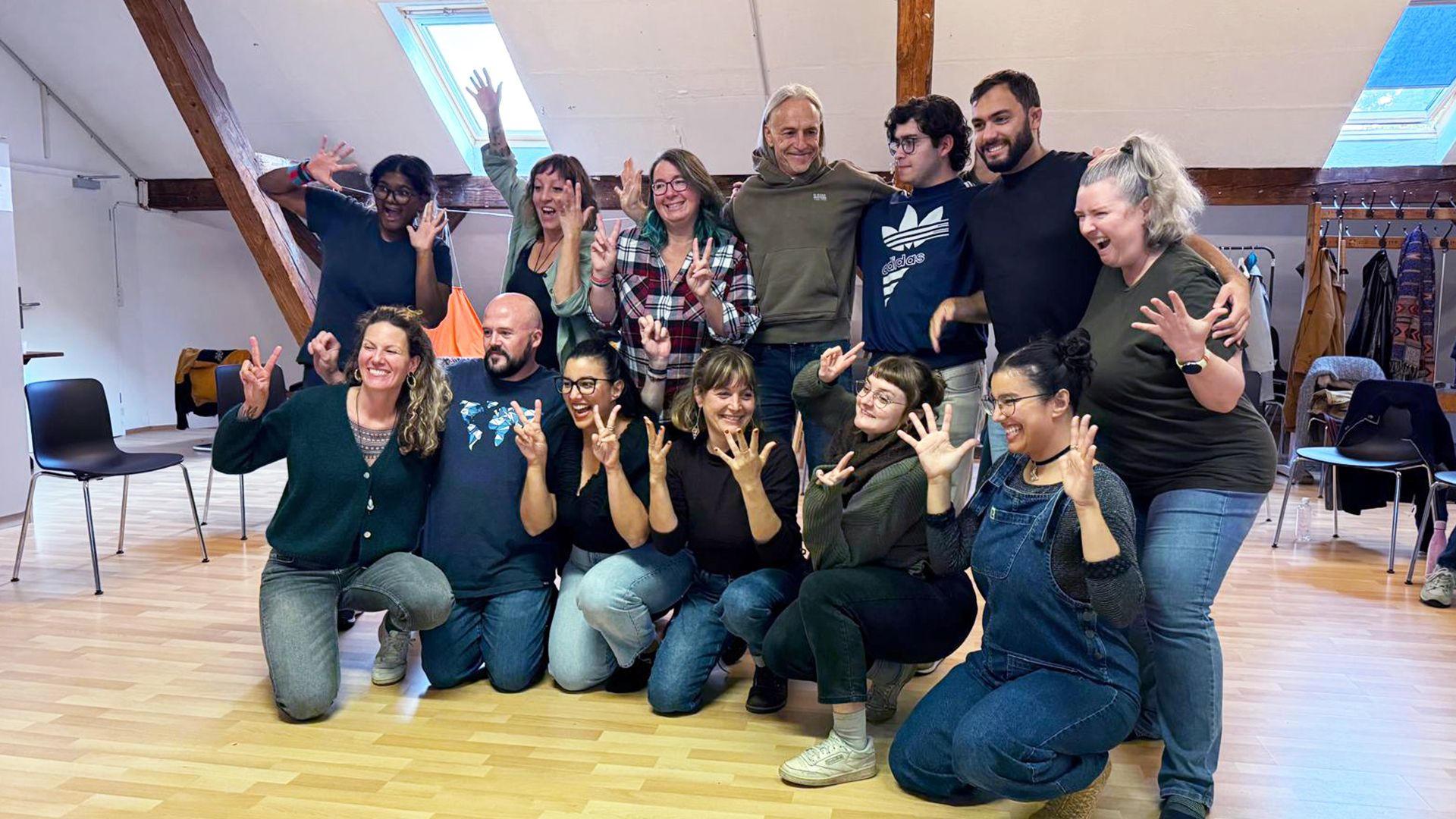 Auf dem Foto sind Amina und Jamila Ouahid mit den 9 Teilnehmerinnen und den 4 Teilnehmern. Alle blicken an der Kamera vorbei, lächeln gut gelaunt und haben ihre Hände in die Höhe gehoben. Ein paar bilden mit dem Zeige- und Mittelfinger ein V, die Gebärde für Visual Vernacular. 