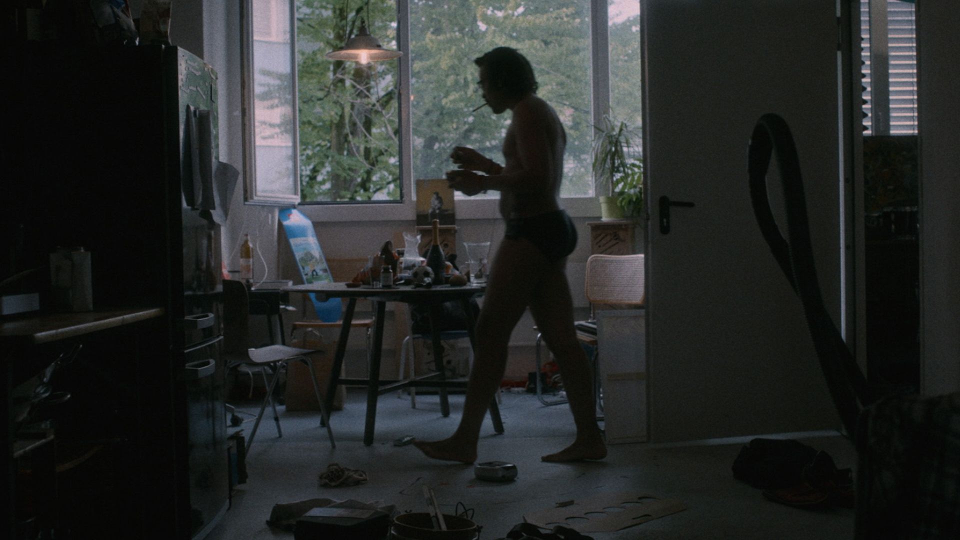 Der Film macht psychische Erkrankung auf eine unverblümte Art und Weise sichtbar macht. Dino Brandão in seinem Studio. Bildnachweis: Outside the Box.
Dino Brandão läuft mit Zigarette im Mund und in Unterwäsche durch sein schwach beleuchtetes Studio. Es liegen Aschenbecher, Kartons und viele andere Sachen herum.