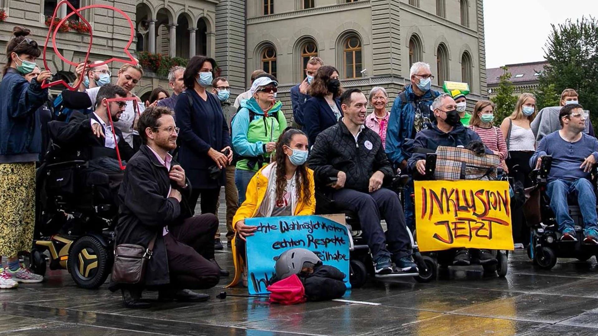Eine Gruppe von ca. 20 Menschen steht vor dem Bundeshaus, dessen Sandsteinmauern im Hintergrund zu sehen sind. Alle blicken in Richtung des rechten Bildrandes, vermutlich in Richtung einer anderen Kamera. Es gibt verschiedene bekannte Gesichter der Behindertenbewegung: Islam Alijaj, Tatjana Binggeli, und Cyril Mizrahi und weitere. Viele tragen eine Maske, was einen Zusammenhang mit der damals akuten Covid-Pandemie haben dürfte. Eine Person im Rollstuhl hat zu ihren Füssen ein grosses gelbes Kartonschild platziert:«Inklusion jetzt» steht drauf.