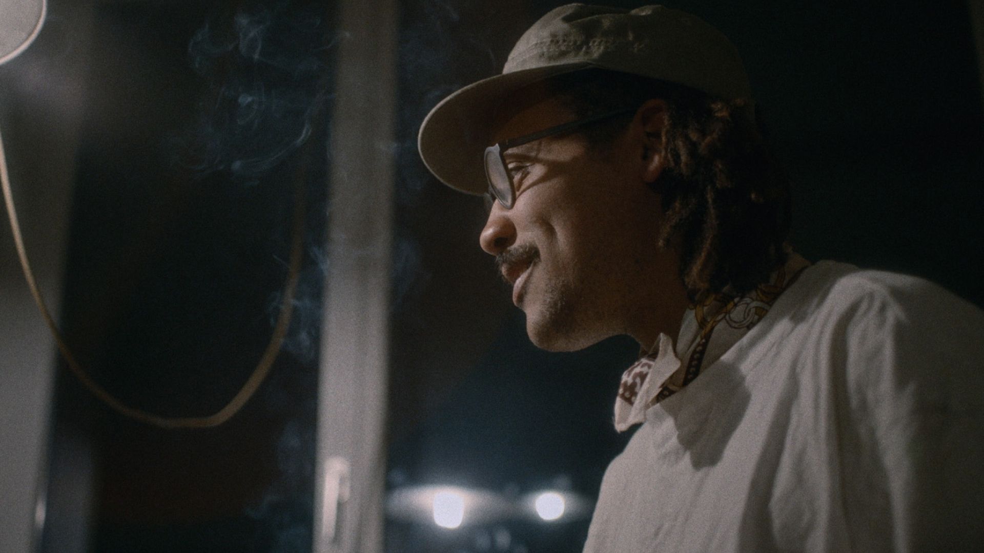 Der Film begleitet Musiker Dino Brandão und sein Umfeld während einer manischen Episode seiner bipolaren Erkrankung. Bildnachweis: Outside the Box.
Dino Brandão in seitlicher Ansicht steht in einem schwach beleuchteten Raum, trägt eine helle Kappe, Brille und ein weisses Hemd. Leichter Rauch steigt neben ihm auf, während warmes Licht sein Gesicht hervorhebt.