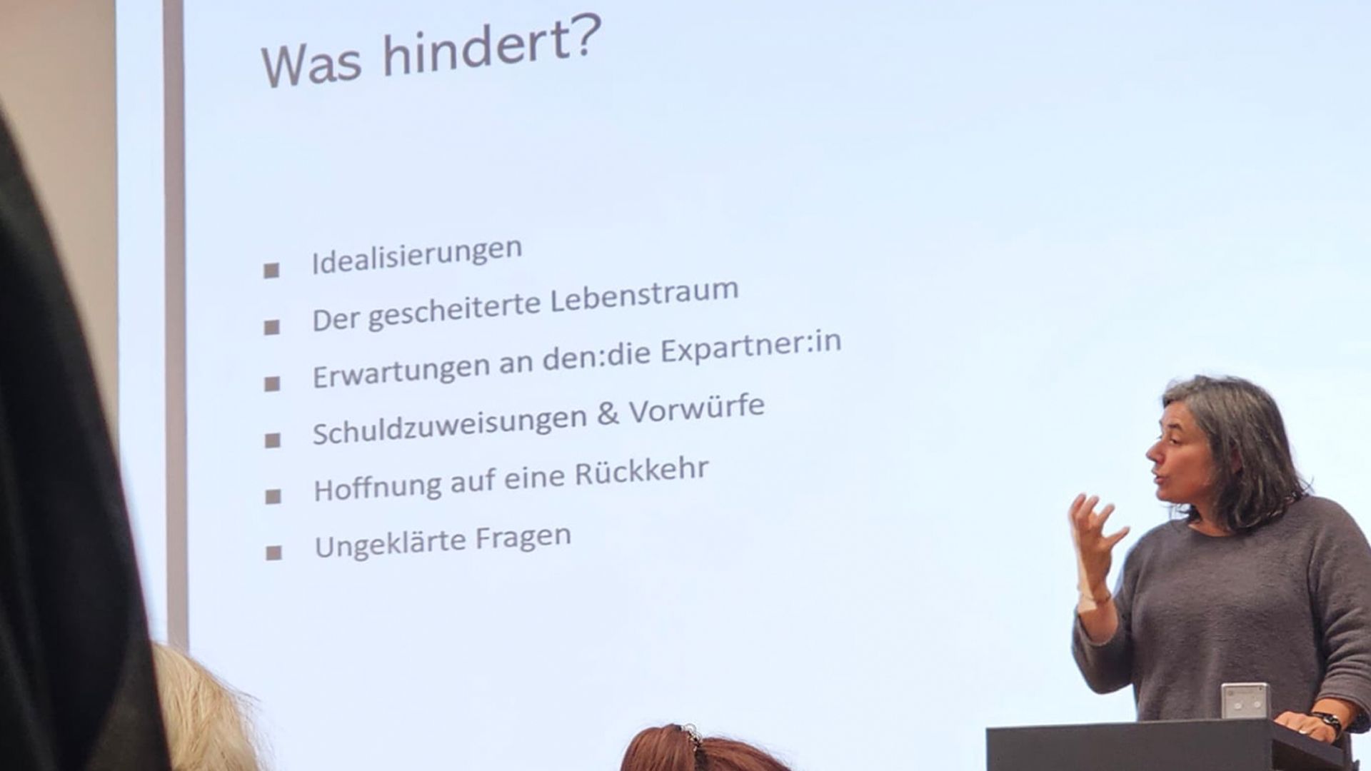 Annette Cina steht hinter einem Rednerpult. Hinter ihr wird gross eine Präsentation an die Wand projeziert.