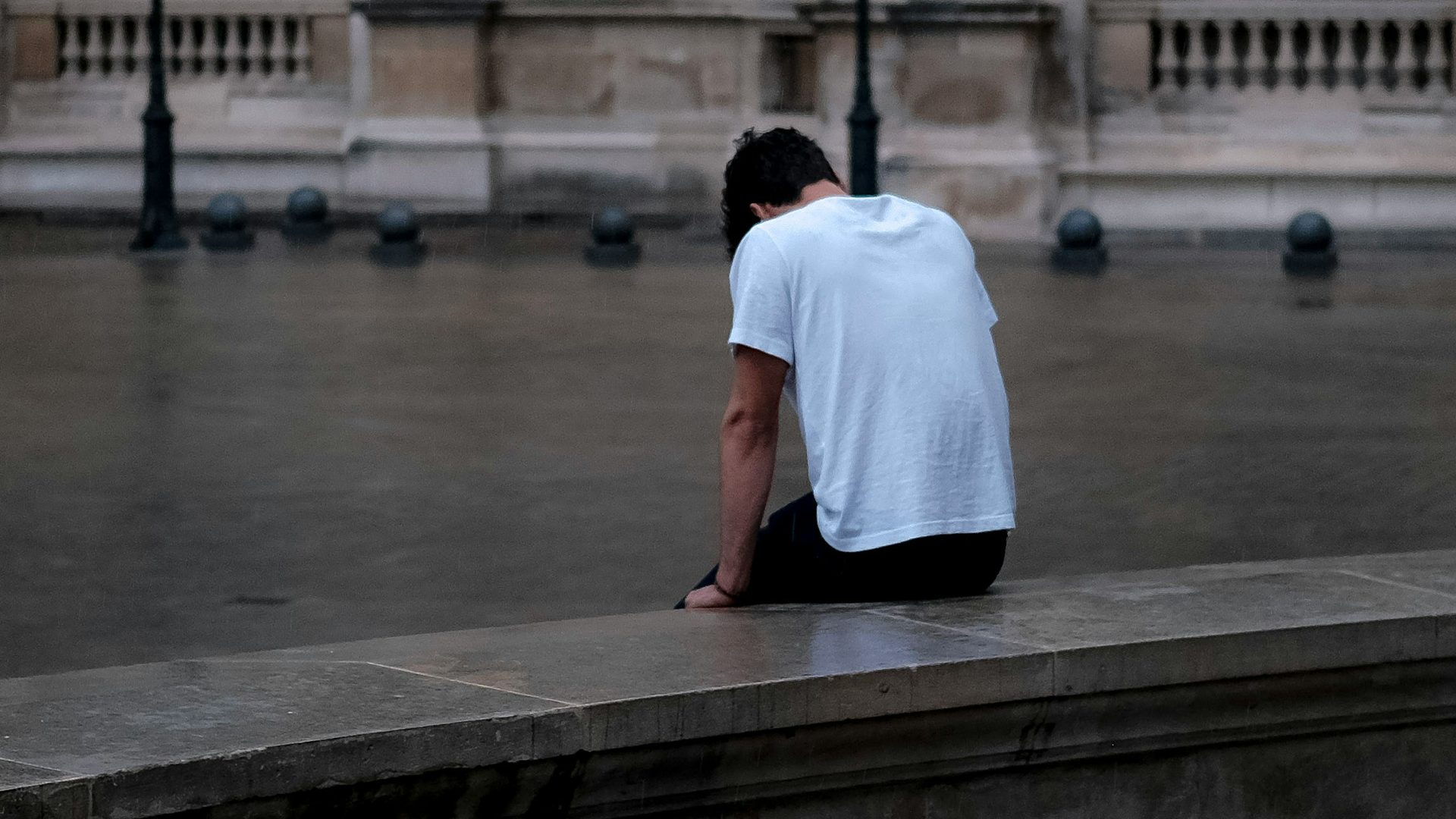 Ein Mann trägt ein weisses T-Shirt und sitzt draussen auf einer Ablage. Er hat den Rücken zur Kamera gedreht. Die Hände stützt er zur Seite ab. Er lässt den Kopf hängen.