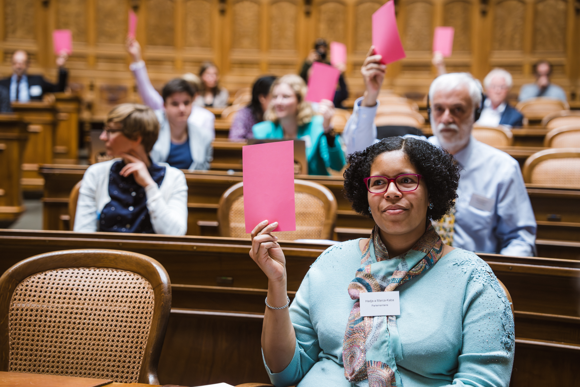 Parlamentarier:innen der Behindertensession halten bei einer Abstimmung rosa Karten hoch. Eine Frau im Vordergrund trägt eine Brille und einen Schal. Sie hält ihre Karte hoch.