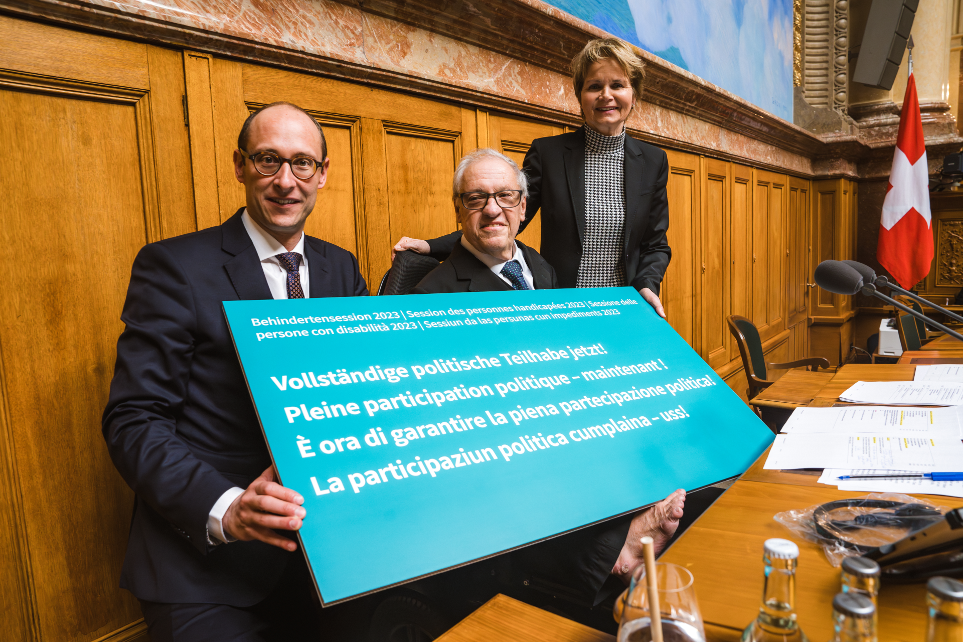 Von links nach rechts: Nationalratspräsident 2023 Martin Candidas, Nationalrat Christian Lohr, Ständeratspräsidentin 2023 Brigitte Häberli-Koller im Nationalratssaal. Sie halten ein blaues Schild. Darauf steht: "Vollständige politische Teilhabe jetzt!"