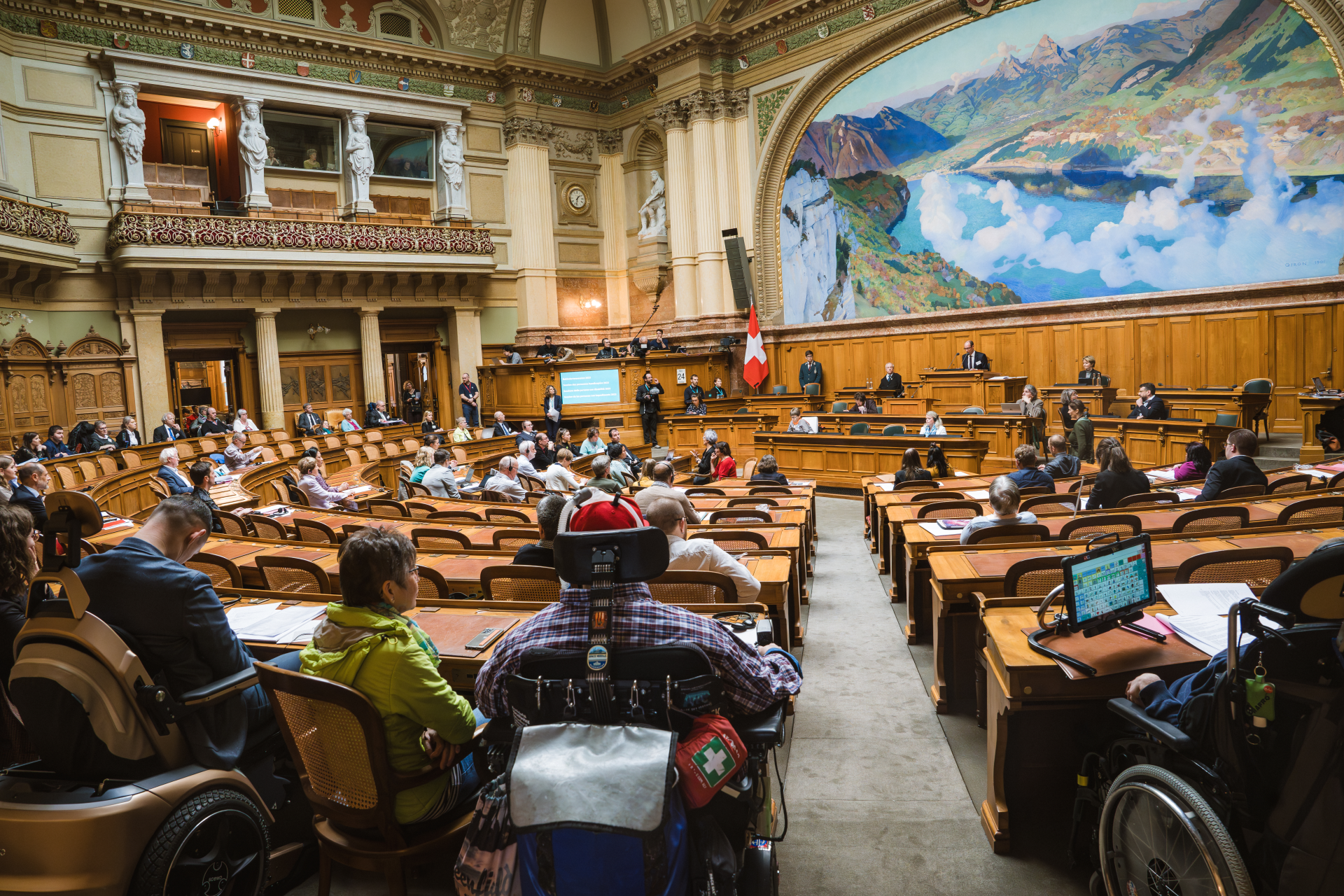 Nationalratsaal mit sitzenden Parlamentarier:innen der Behindertensession. Der Saal hat ein grosses Wandgemälde und eine Schweizer Flagge. Im Vordergrund sind barrierefreie Sitzplätze zu sehen.