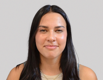 Porträt von Juli Obando. Sie hat lange dunkle Haare, trägt ein helles Oberteil vor einem einfarbigen Hintergrund.