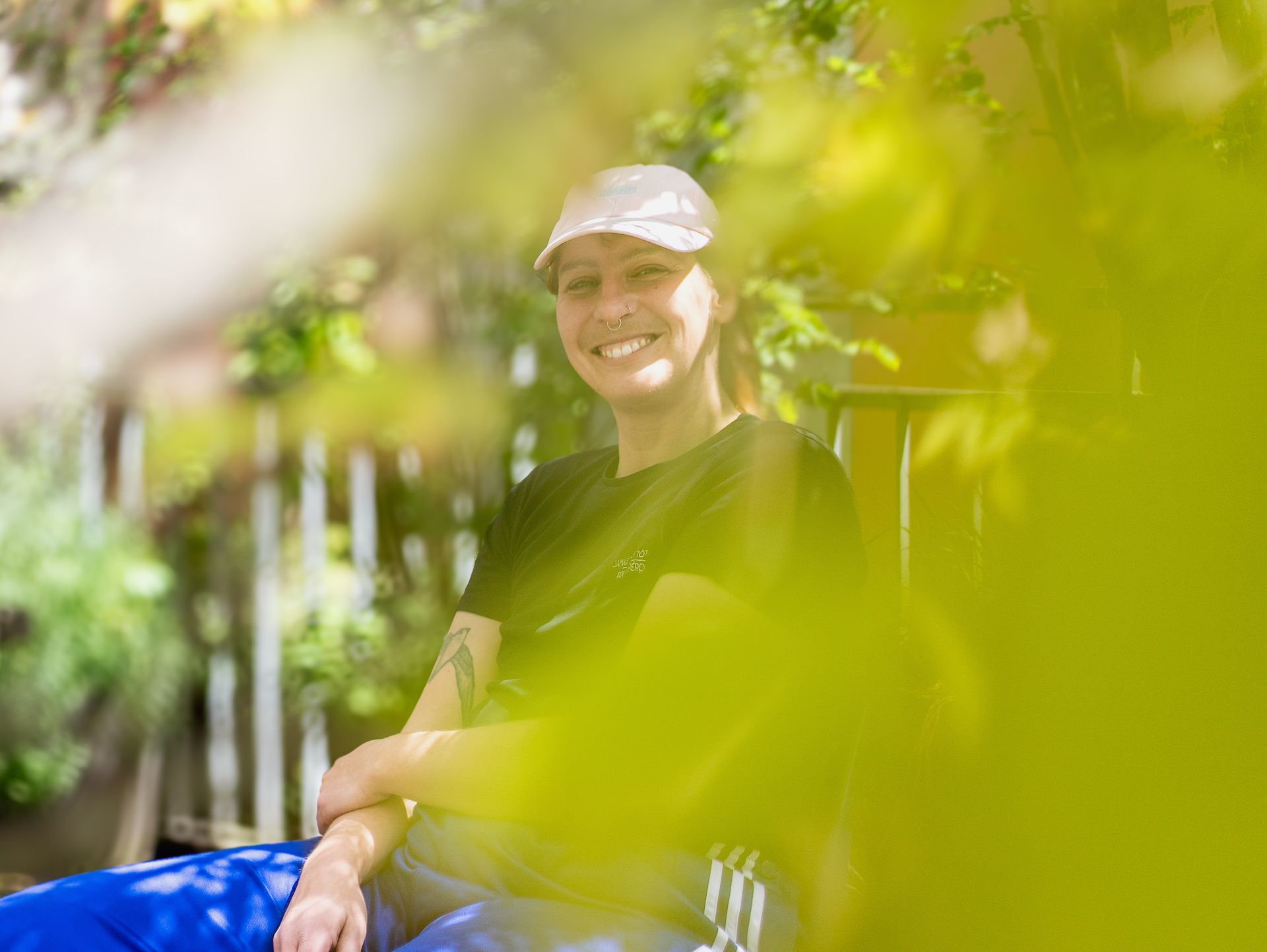 Lovis sitzt und schaut direkt in die Kamera. Lovis lacht. Lovis trägt ein rosa Cap, königsblaue Trainerhosen und ein schwarzes Kurzarmshirt. Am rechten Oberarm ist ein Schwalbentattoo zu sehen. Lovis hat zwei Nasenpiercing-Ringe. Das Foto wurde an einem sonnigen Tag draussen aufgenommen. Lovis sitzt auf einer Terrasse. Es gibt Pflanzen rundherum. Die Aufnahme enthält Unschärfen und wurde durch grüne Äste und Blätter hindurch fotografiert. Dadurch ist Lovis von einem verschwommenen, kunstvoll-abstrakten Grün umrahmt. Bildquelle: Silvia Meierhofer