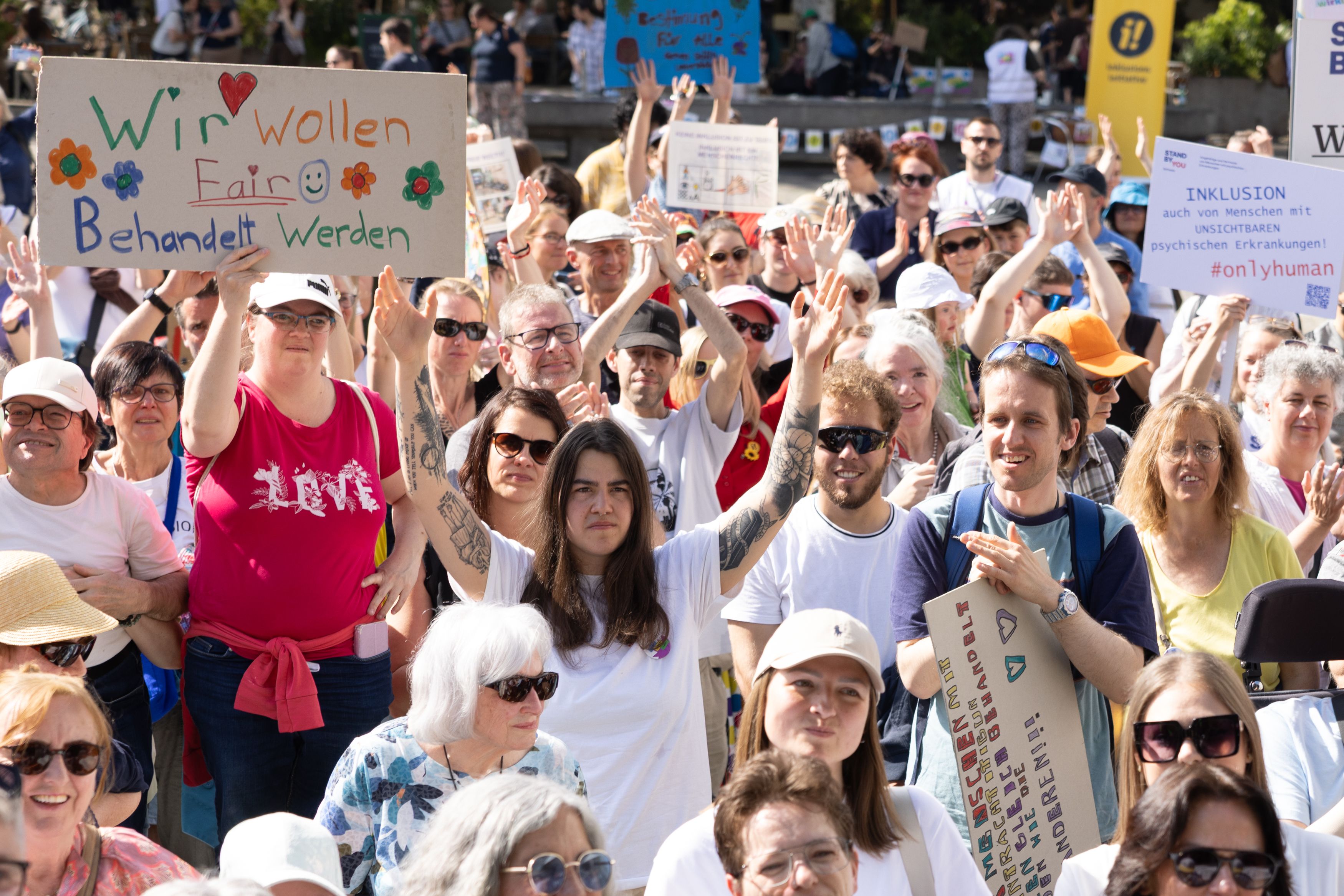 Eine Menschenmenge bei einer Demonstration, die Schilder mit Aufrufen zu fairer Behandlung und Inklusion hochhält; viele tragen Sonnenbrillen und Sommerkleidung.