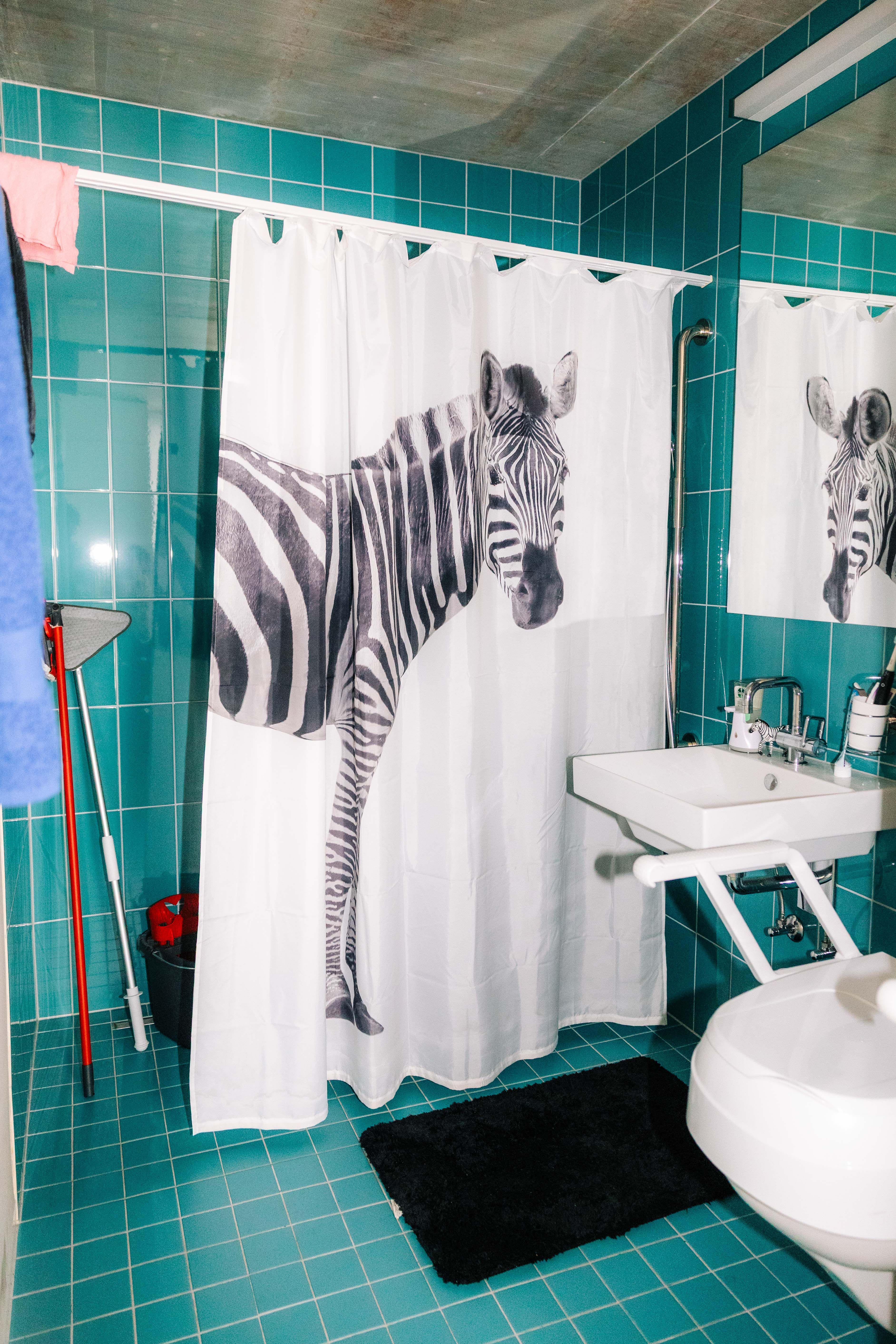 Das Badezimmer von Sandra Kropf ist komplett barrierenfrei. Bild: Surprise/David Fürst Badezimmer mit türkisfarbenen Fliesen, einem Duschvorhang mit Zebradruck, einem weissen Waschbecken, einem Spiegel, Reinigungsmitteln und einer schwarzen Matte.