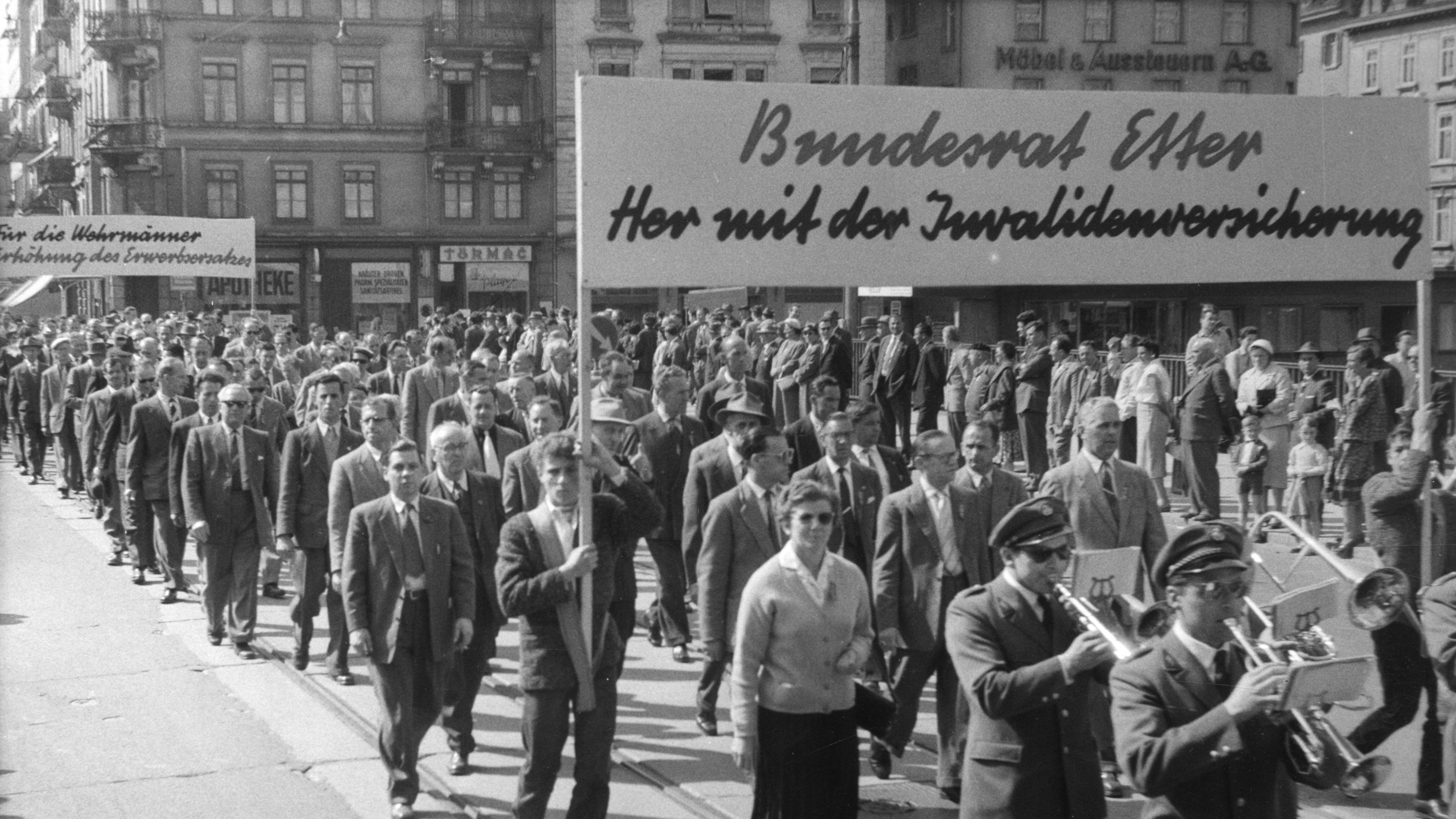 Ein Schwarz-Weiss-Foto einer grossen Menschenmenge, die bei einer Strassenkundgebung marschiert, Schilder mit deutschen Aufschriften hochhält und von einer kleinen Musikkapelle begleitet wird.